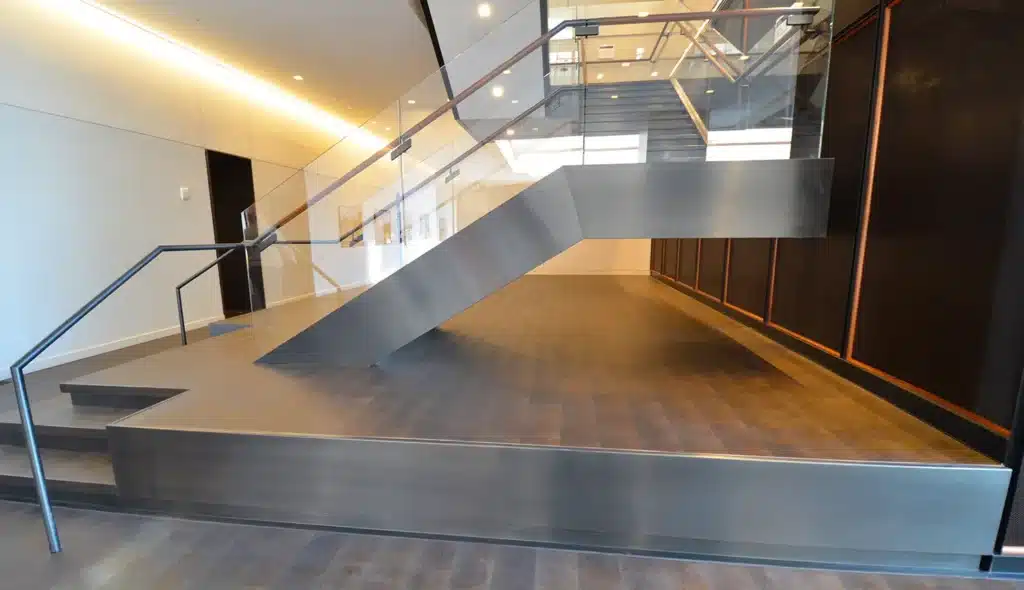 A modern indoor staircase with glass railings and metal supports rises from a raised platform in a sleek, minimalist building lobby with light walls and dark wood flooring.