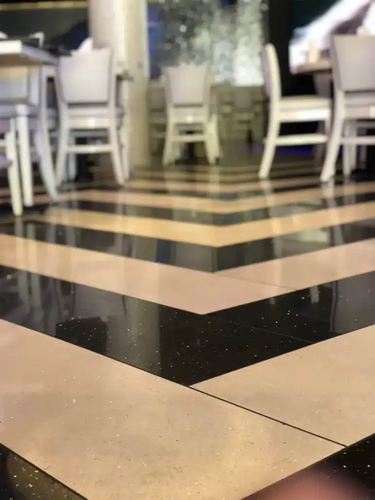 A close-up view of a glossy, black and beige striped floor in a restaurant, with white chairs and tables slightly out of focus in the background.
