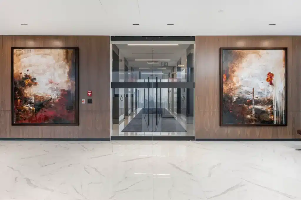 A modern office corridor with large abstract paintings on wood-paneled walls, glossy white marble floor, and glass doors leading to a brightly lit hallway.