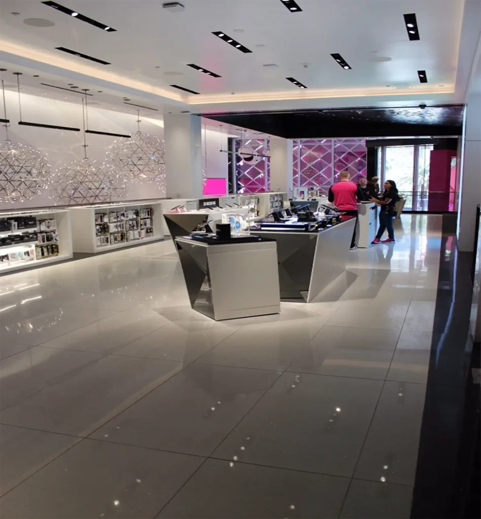 A modern electronics store with sleek white displays and shelves lined with products. Three people stand near the entrance talking, while the store floor and lighting create a bright, spacious atmosphere.