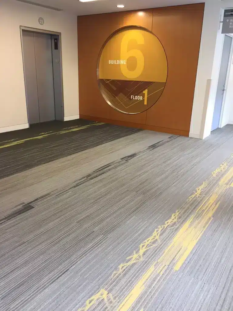 A modern hallway with gray and yellow carpet, an elevator on the left, and a large circular wall sign reading 
