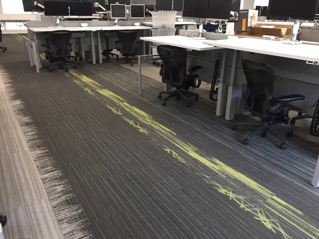 Modern office with several empty desks and ergonomic chairs. The carpet is mostly gray with a noticeable, irregular yellow streak running through the walkway area. Multiple monitors are on the desks.