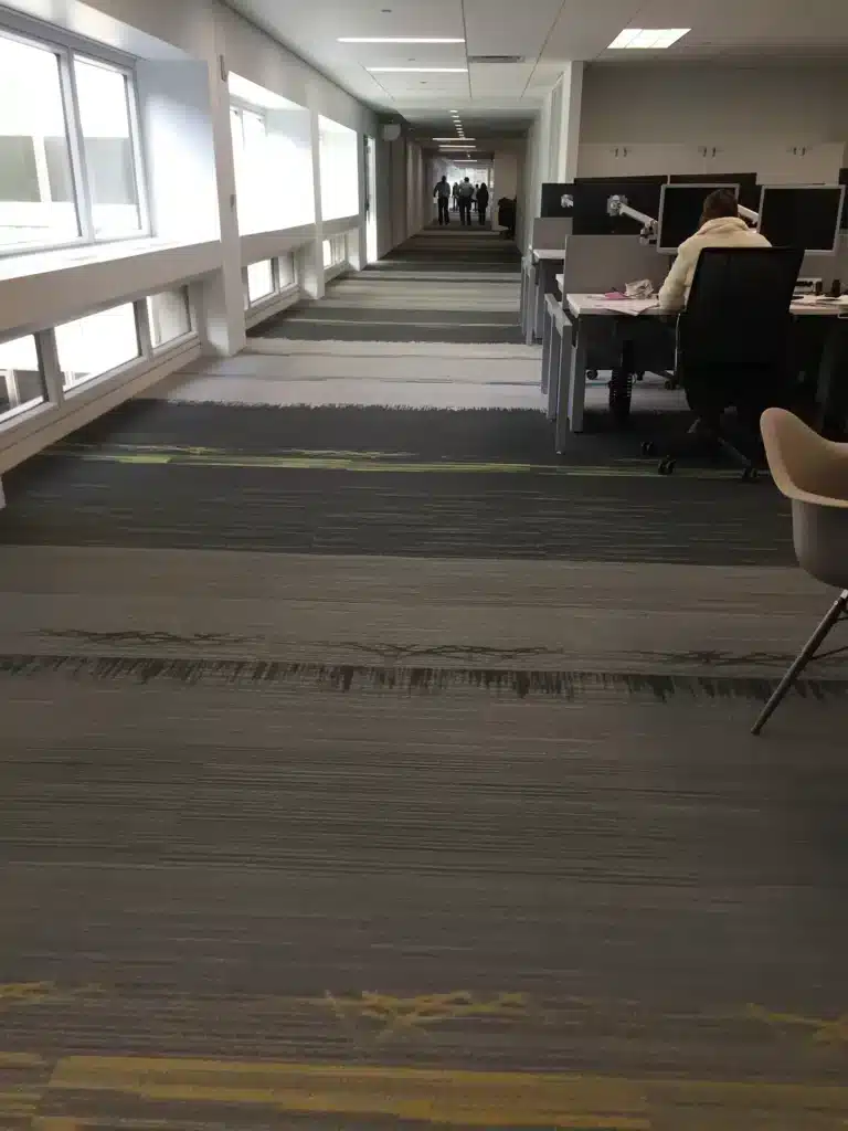 A long office hallway with windows on the left, desks and chairs on the right, and several people walking in the distance at the far end of the corridor. One person is seated and working at a desk.