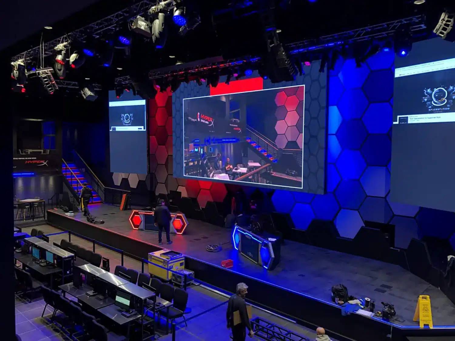 A large esports stage with blue and red hexagonal backdrop lighting, big screens displaying game graphics, several desks with monitors, and a few people setting up equipment. A caution sign is on the far right.