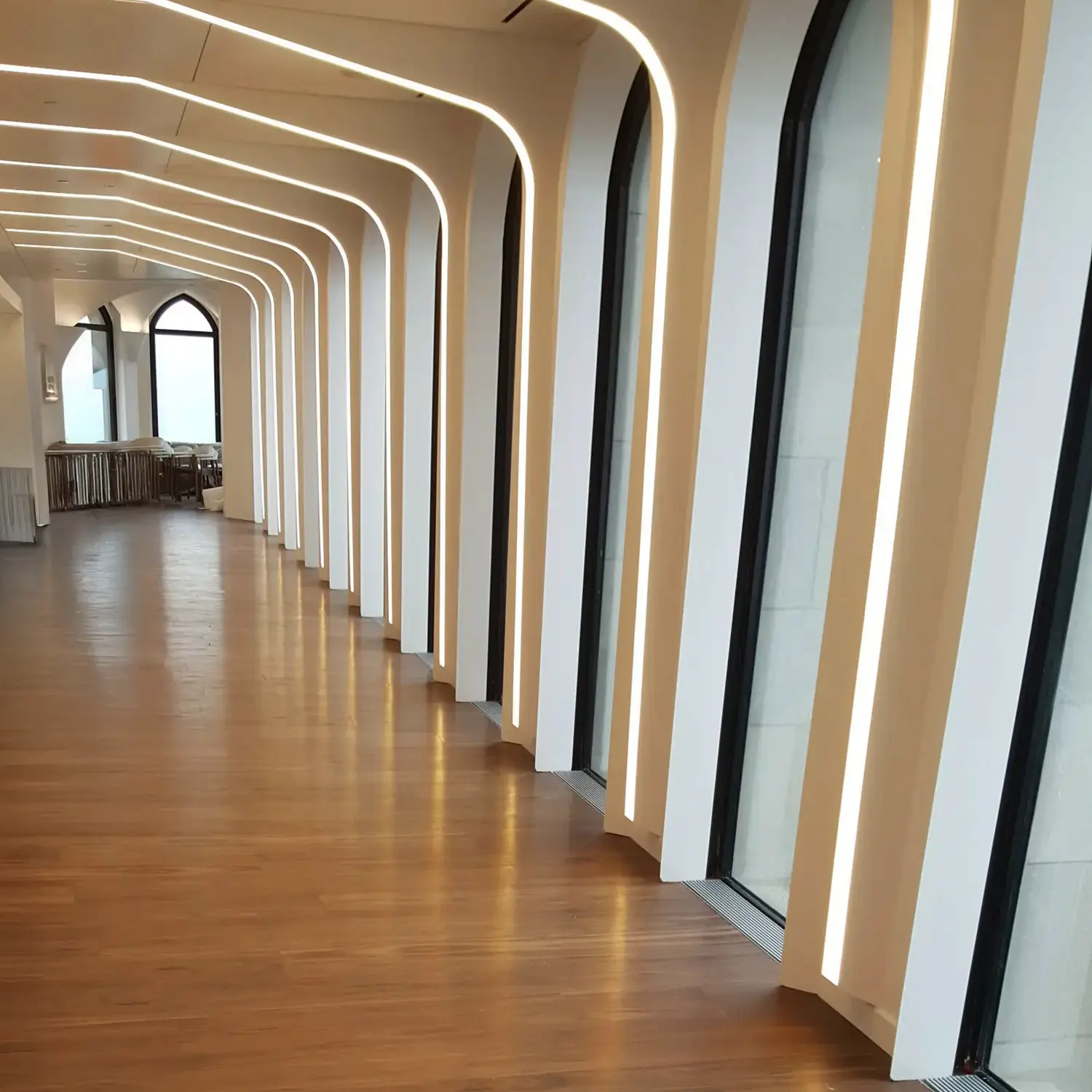 A modern hallway with tall, arched windows and vertical LED light strips along white frames. The floor is wood, and the corridor has a bright, minimalist design with clean lines.