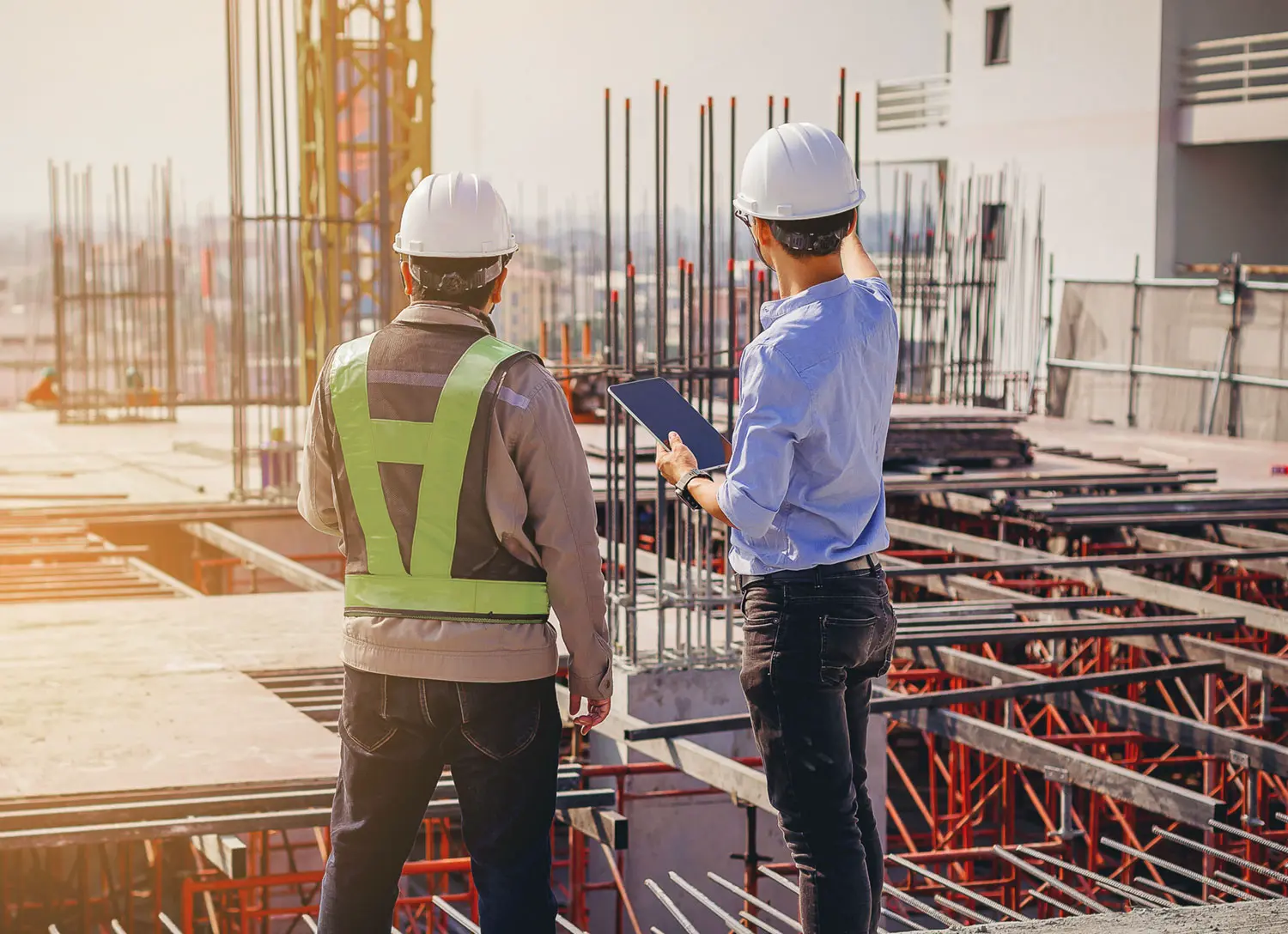 Two construction workers wearing safety gear and helmets stand on a building site with steel reinforcements; one holds a tablet while the other listens, as they discuss the ongoing construction project.