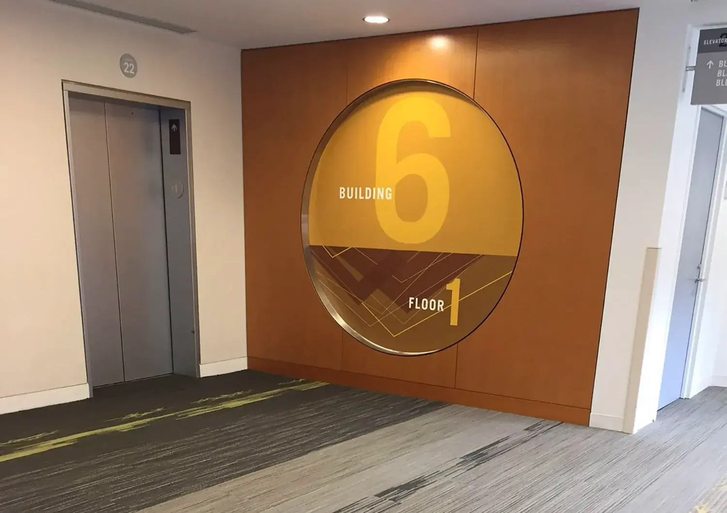 An elevator lobby with a large circular sign on a wooden wall that reads "Building 6, Floor 1" in yellow letters. The elevator doors are closed, and the floor has a modern pattern.