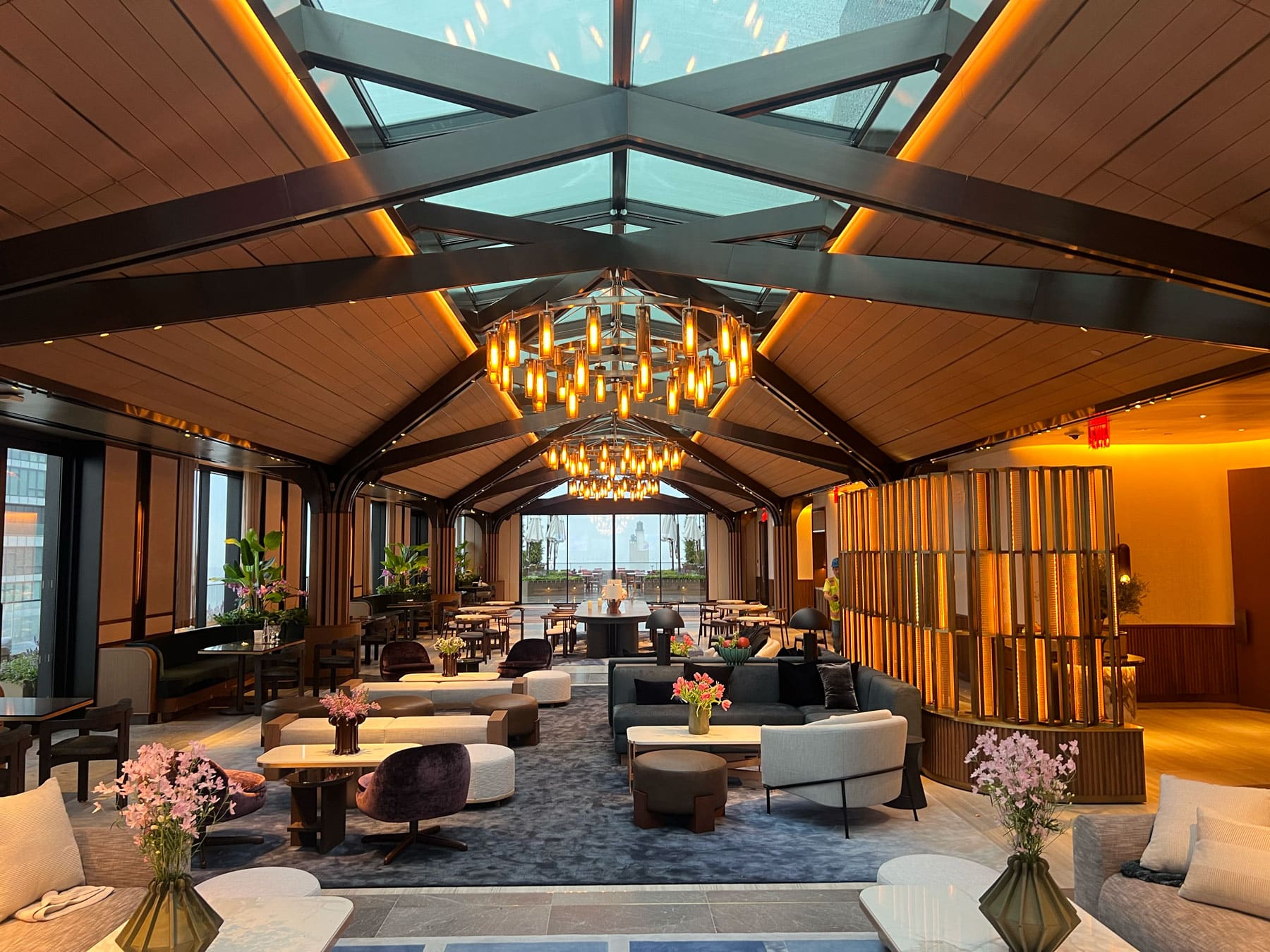 A spacious, modern lounge with a glass ceiling, chandeliers, cozy seating areas, tables with flower arrangements, and large windows at the far end letting in natural light.