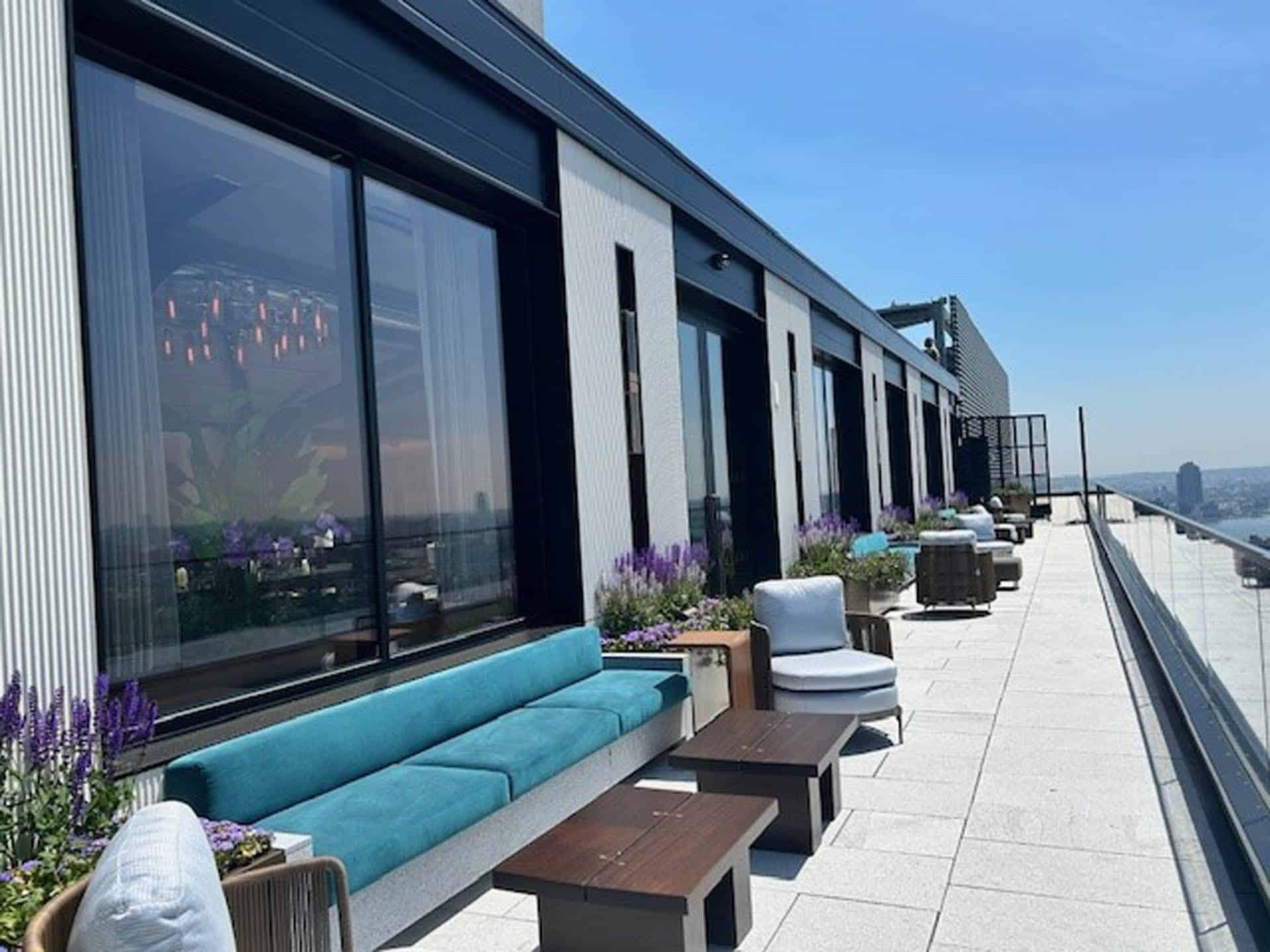 A modern rooftop terrace with blue cushioned seating, tables, and lounge chairs along a glass railing, decorated with potted purple flowers and overlooking a city skyline on a sunny day.