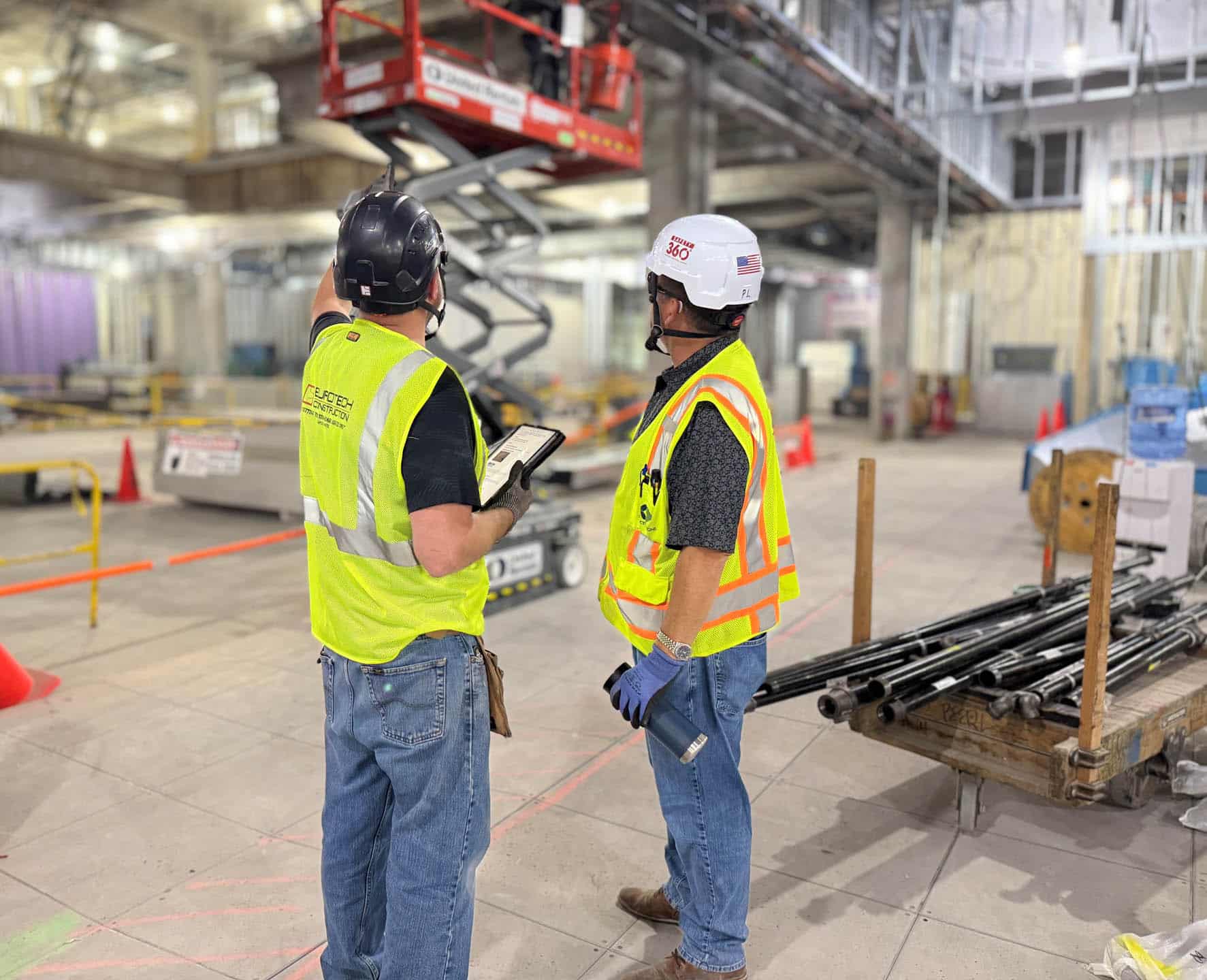 Two construction workers in safety vests and helmets stand in a large, unfinished building. One points towards a worker on a lift while the other listens and holds a flashlight. Scattered materials and equipment are visible.