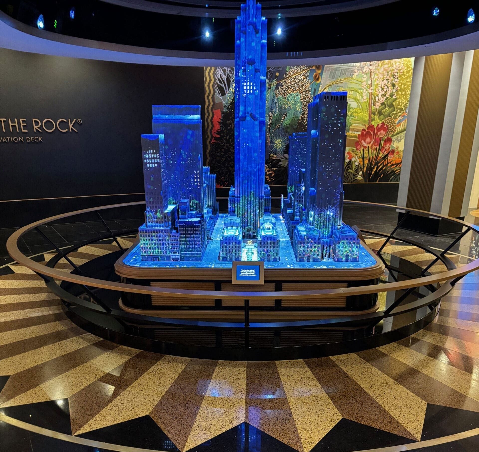 A detailed, illuminated scale model of New York City skyscrapers, featuring blue lighting, is displayed on a round platform indoors. The background includes a vibrant floral mural and polished patterned flooring.