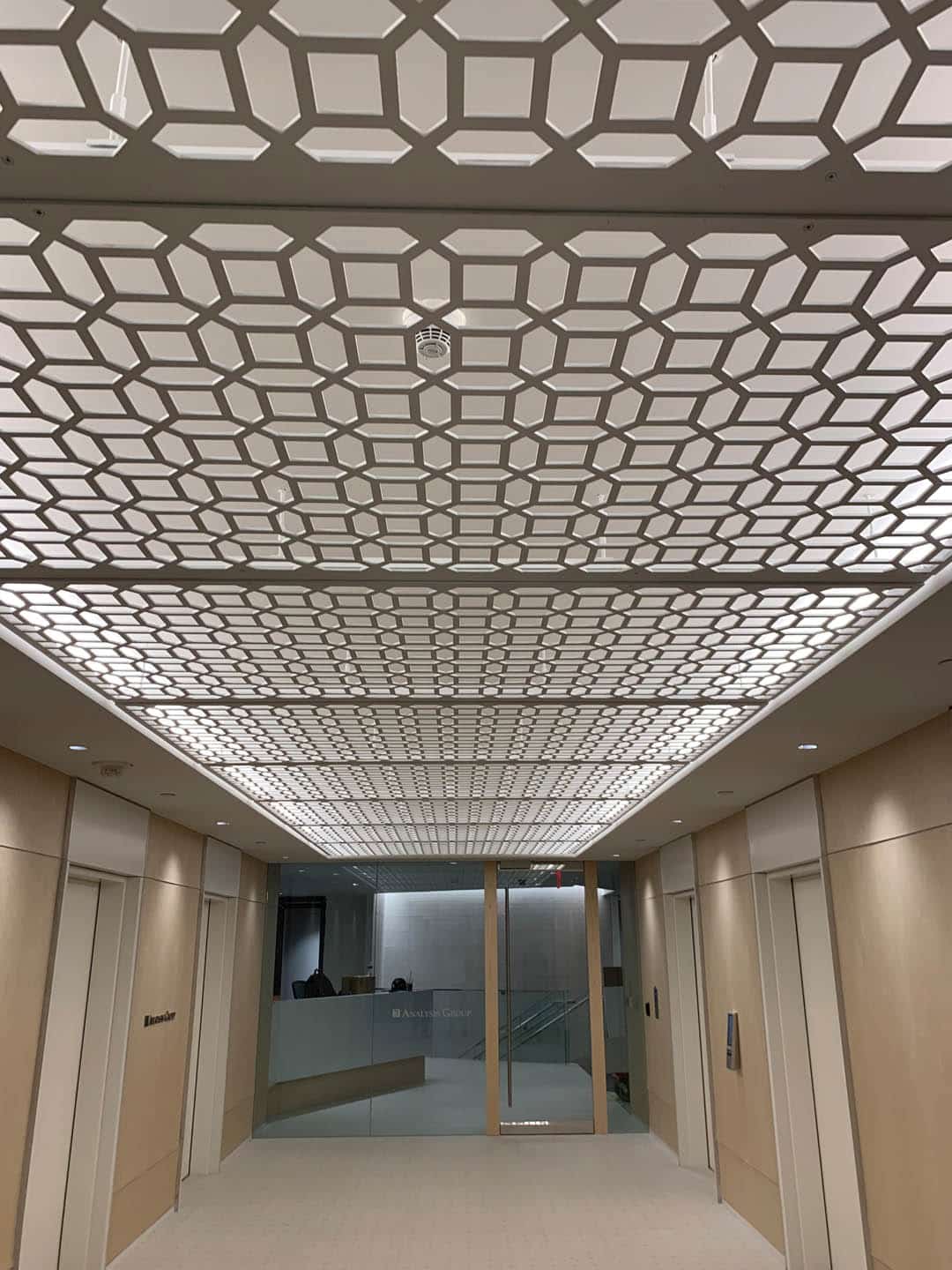 Modern hallway with light wood walls and elevators on each side. The ceiling features an intricate geometric pattern with bright lighting, leading to a reception desk and glass doors at the end of the corridor.