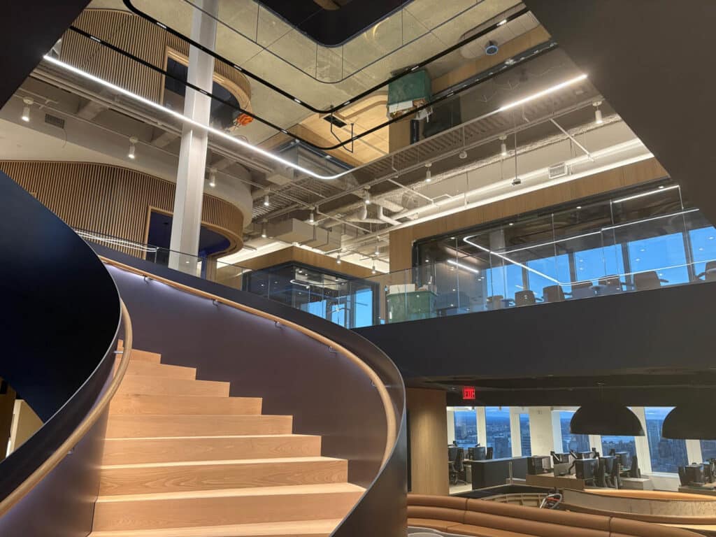 A modern office interior with a curved wooden staircase, open seating areas, glass-walled meeting rooms, and large windows letting in natural light from outside.
