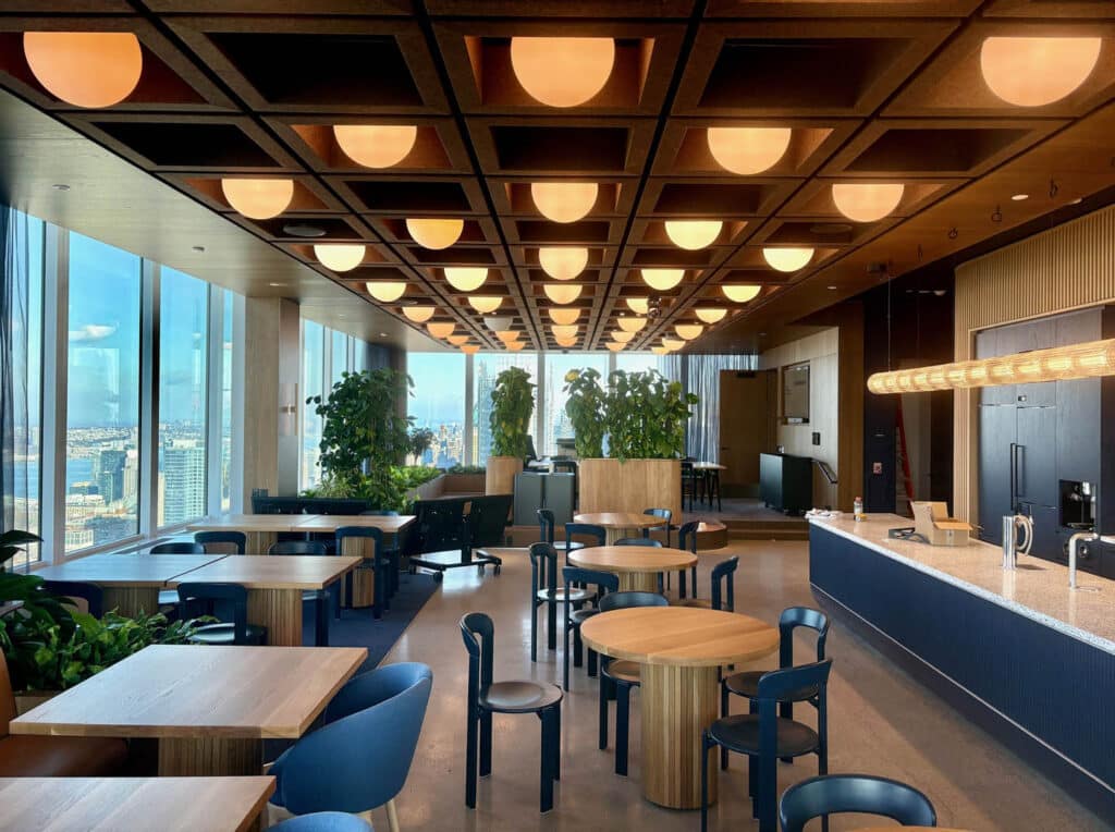 Modern office lounge with round and square tables, black chairs, large windows with city views, indoor plants, and a wooden ceiling with geometric lighting. A counter with coffee machines is on the right.
