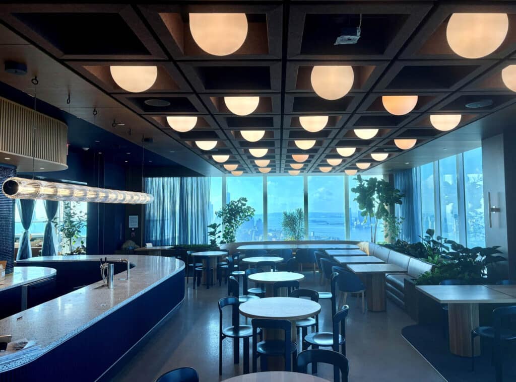A modern lounge with round tables, blue chairs, a curved bar, and large windows showcasing an ocean view. Ceiling lights are evenly spaced, and potted plants decorate the space.