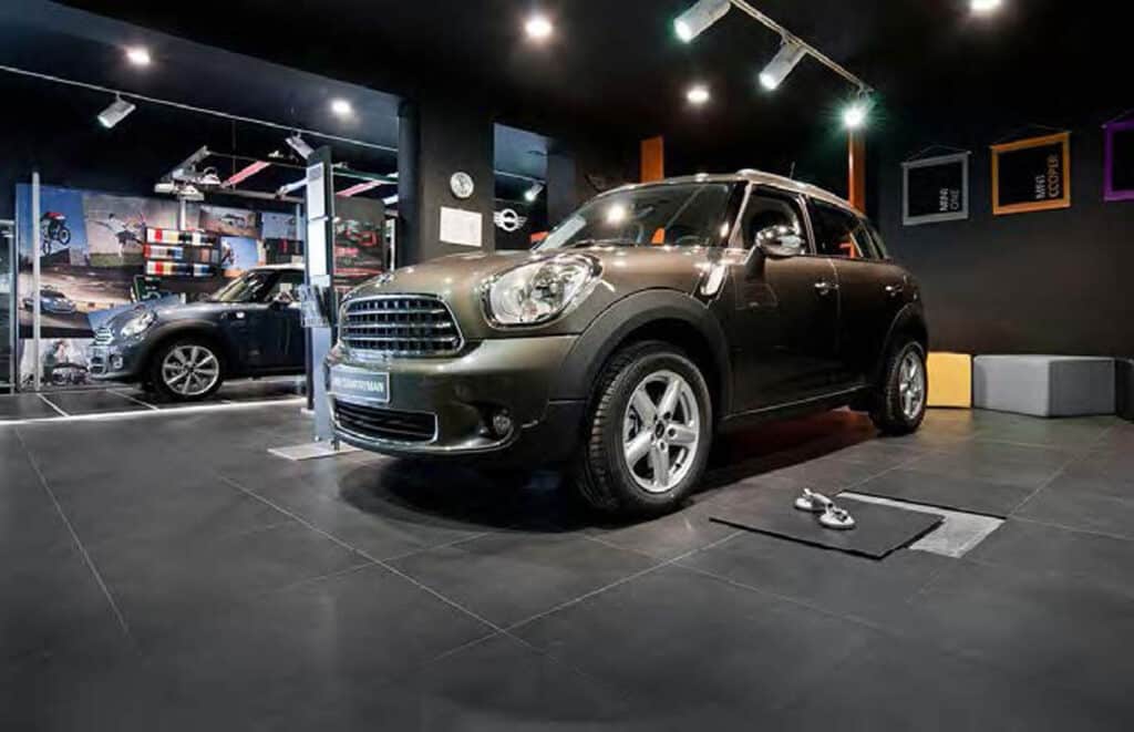 A black Mini Cooper is parked indoors on a tiled floor next to a mat with two dumbbells. The room has dark walls, modern decor, and colorful cube seats in the background.