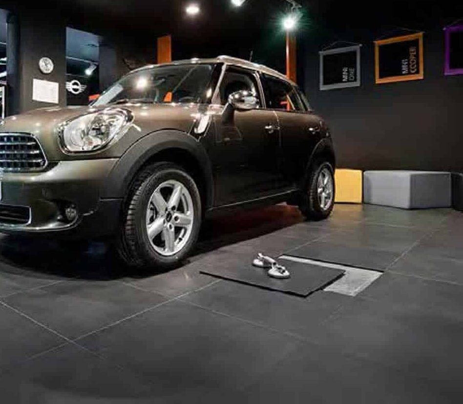 A black Mini Cooper is parked indoors on a tiled floor next to a mat with two dumbbells. The room has dark walls, modern decor, and colorful cube seats in the background.