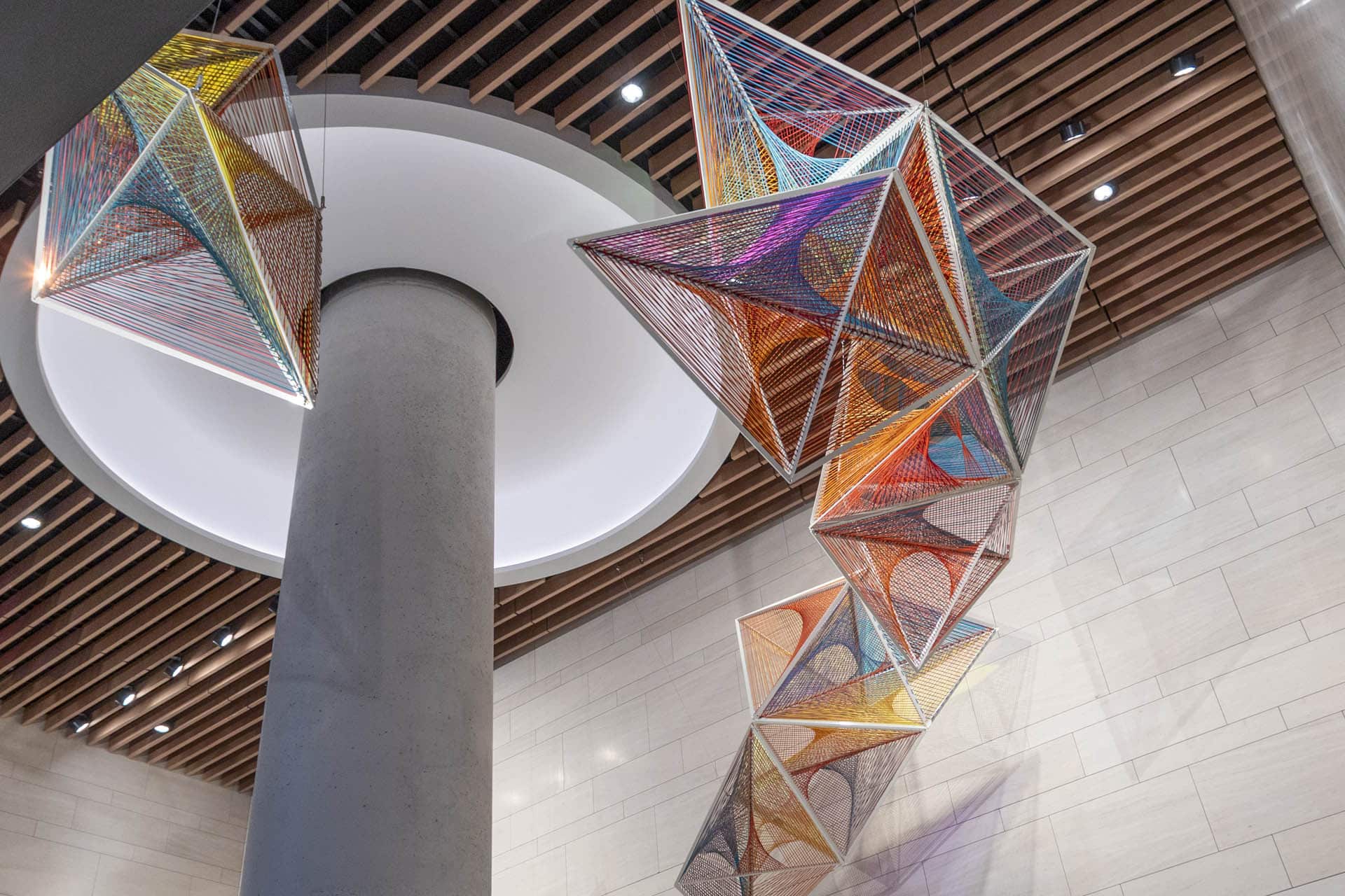 Colorful geometric sculptures made of strings form interconnected pyramids and cubes, hanging from the ceiling near a circular skylight and a column in a modern building lobby with wood and stone details.