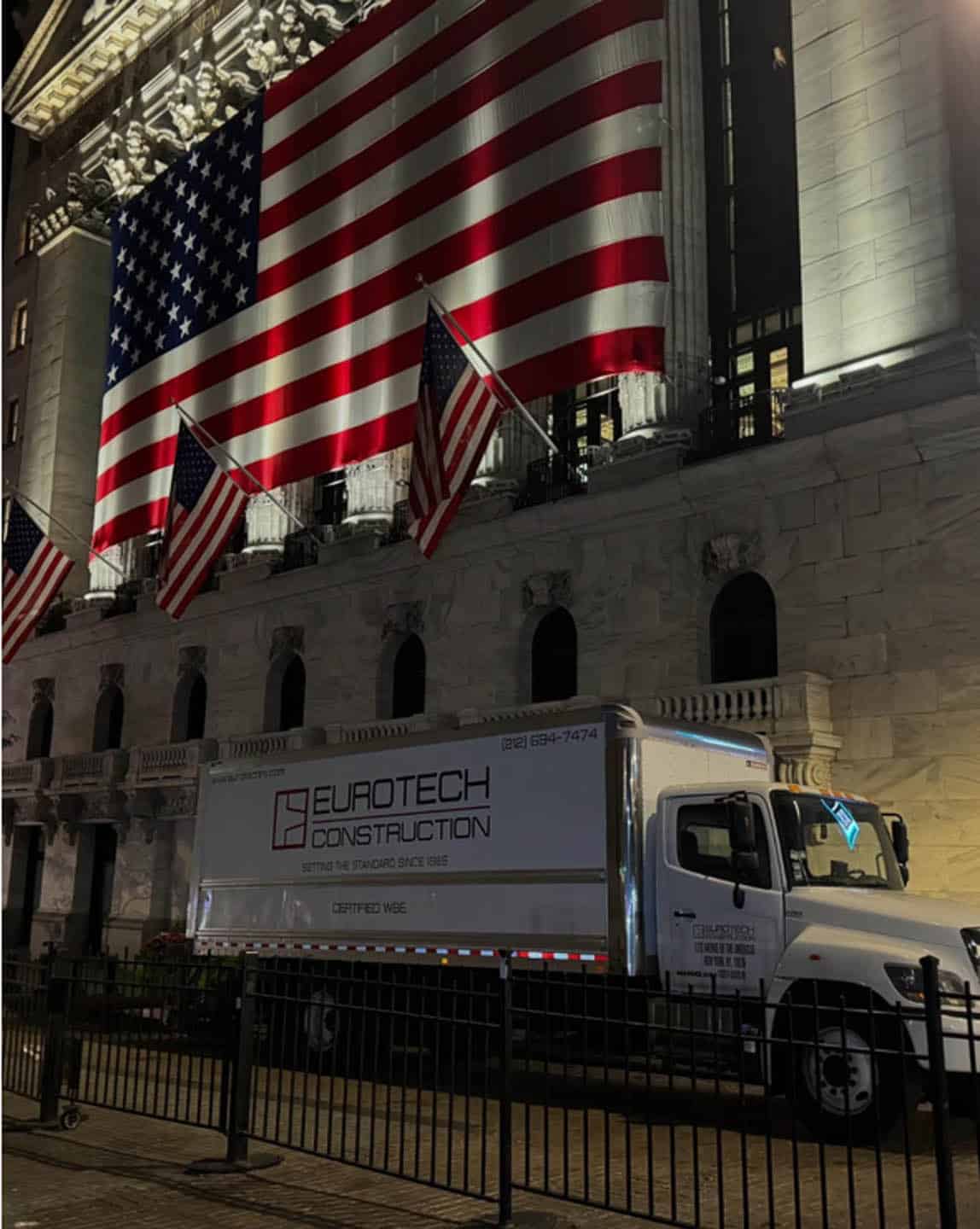 A large American flag and two smaller flags hang on the facade of a building at night. A Eurotech Construction truck is parked in front of the building, which has stone walls and is illuminated by spotlights.