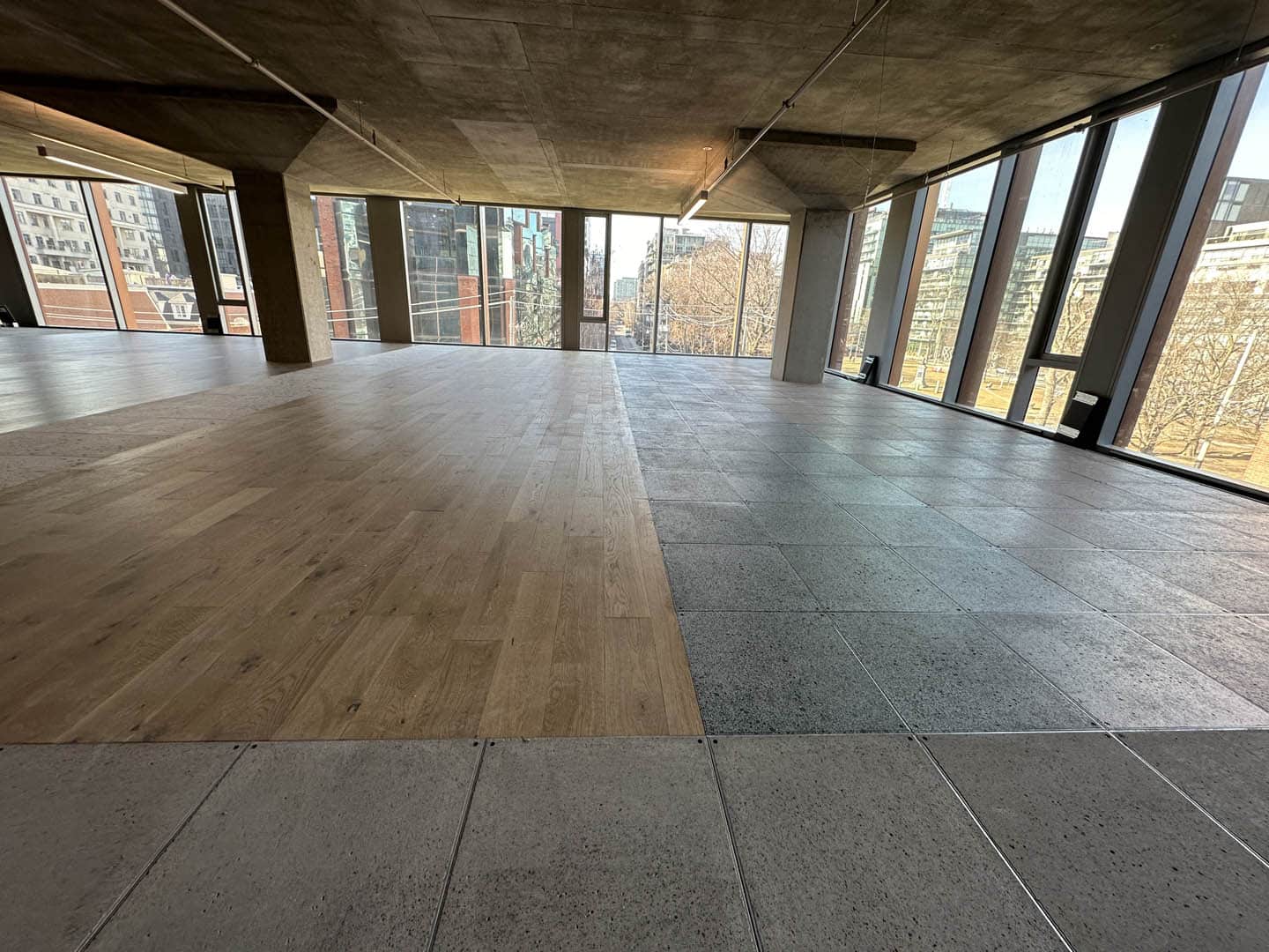 A spacious, empty room with large floor-to-ceiling windows. The floor is divided into two sections: one with light wood and the other with gray tiles. City buildings and trees are visible outside.