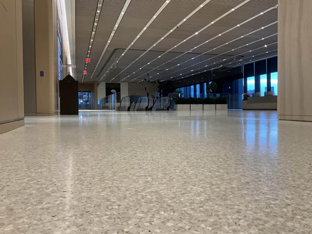 A modern, empty building lobby with polished terrazzo floors, overhead lights, wood-paneled walls, and escalators leading to an upper level in the background. Large windows show a view of the outside.