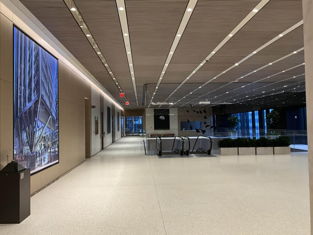A spacious, modern building lobby with a light-colored floor, large architectural artwork on the wall, an escalator in the center, ceiling lights, and floor-to-ceiling windows letting in natural light.