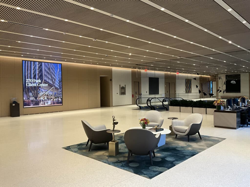 Spacious modern lobby with a seating area of four gray chairs around a small table on a blue rug, large wall art, an escalator, and a reception desk in the background.