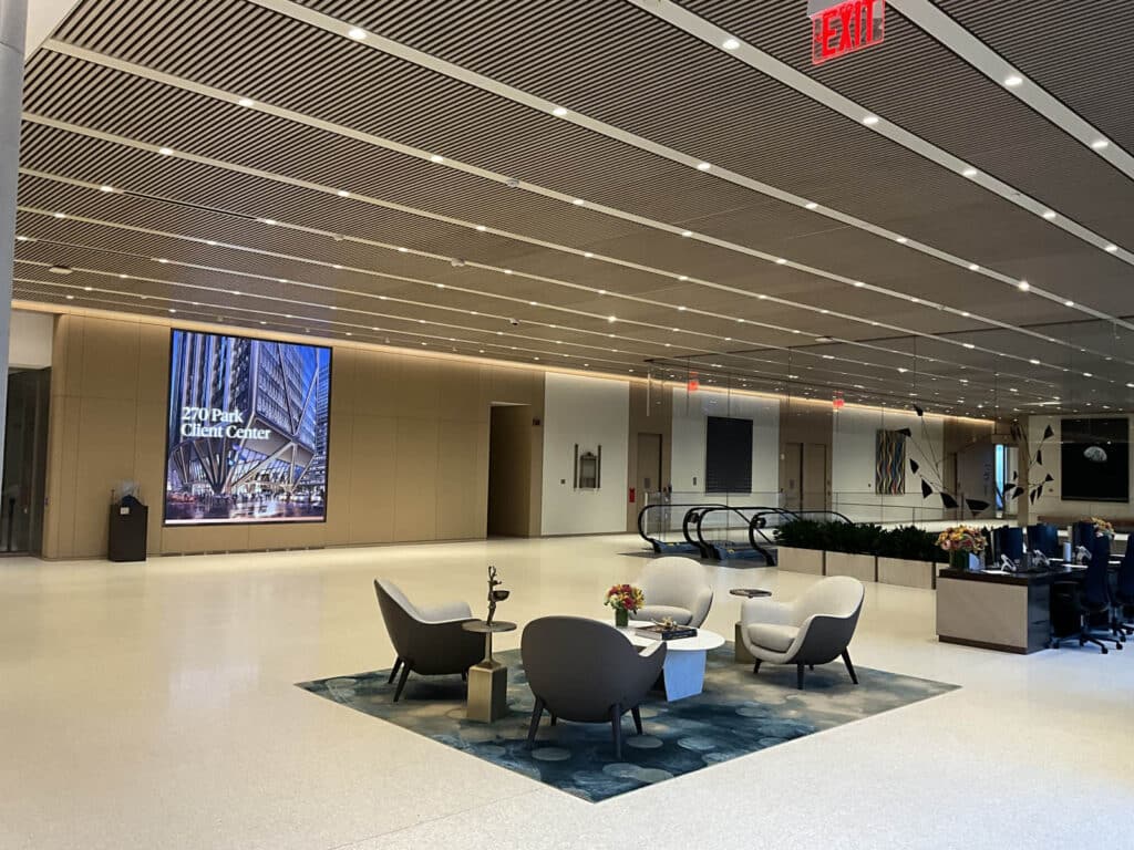 Modern office lobby with seating area, four chairs around a small table on a rug, large digital display on the wall, escalators, workstations, and bright overhead lighting.
