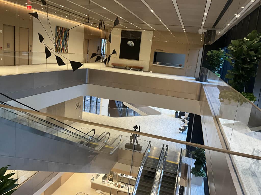 Modern multi-level building interior with escalators, glass railings, abstract art on walls, benches, and large potted plants. Clean, open space with natural light and sleek architectural design.