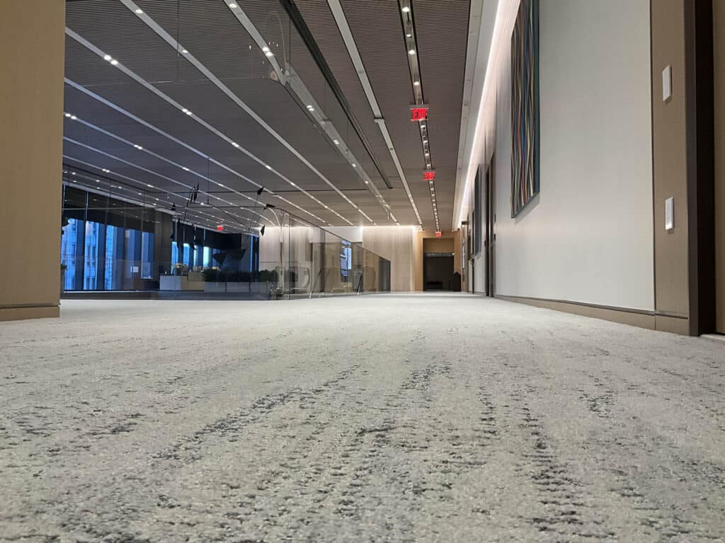 Low-angle view of a modern office hallway with a carpeted floor, glass walls, wooden panels, ceiling lights, and abstract art on the wall; office furniture visible in the background.