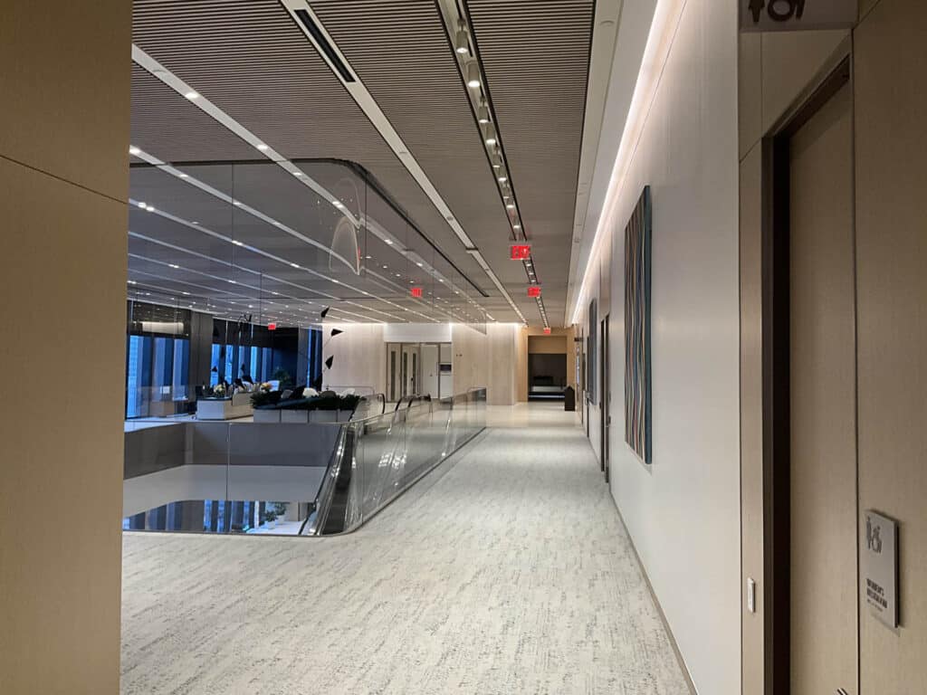 A modern, well-lit office hallway with carpeted flooring, glass railings, and large windows. Artworks hang on the right wall, and there is a door with a sign on the right. Red exit signs are visible on the ceiling.