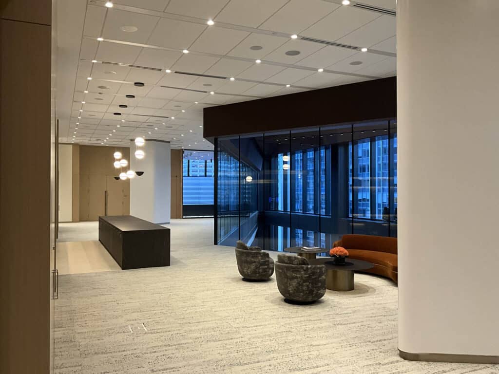 Modern office lobby with large windows, a long dark reception desk, two patterned armchairs around a low table, and an orange sofa. The space is well-lit with ceiling and pendant lights.