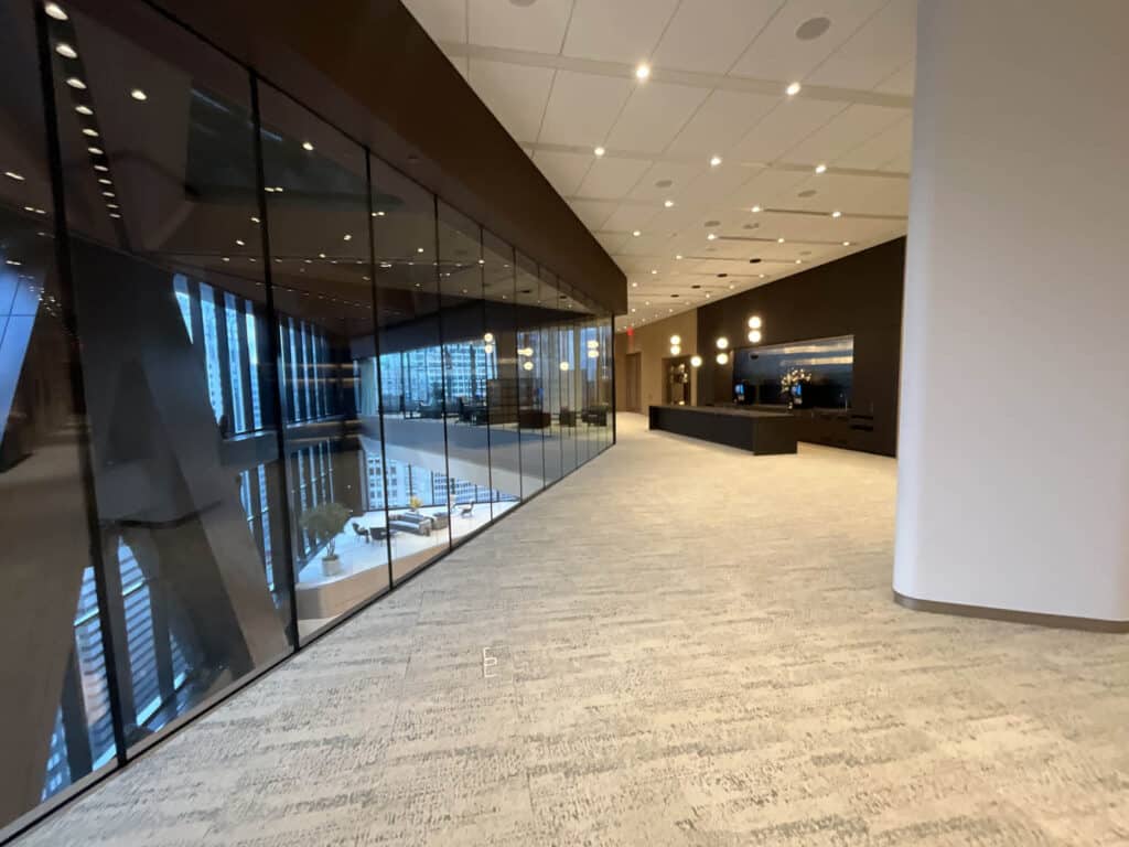 Spacious modern office lobby with large windows, beige carpet, and dark accent walls. Reflections show tall city buildings outside. There is a reception desk and decorative lights on the far end.