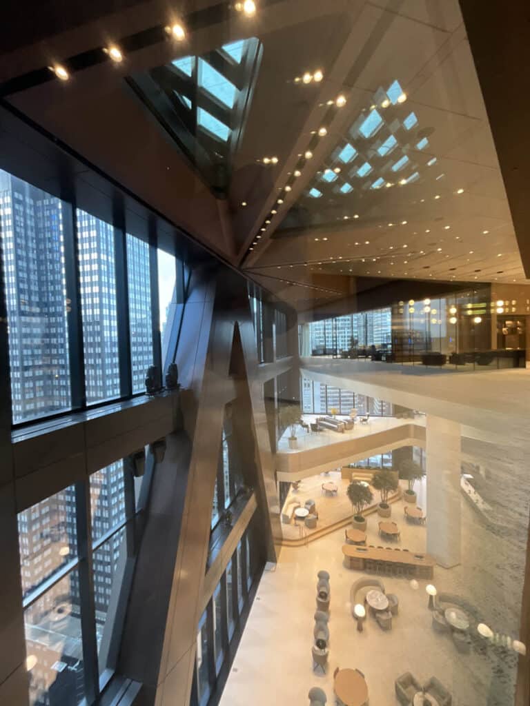 Modern office building interior with large glass windows, geometric steel beams, and a spacious lobby area with seating and plants. City skyscrapers are visible through the windows, and ceiling lights reflect off the glass.