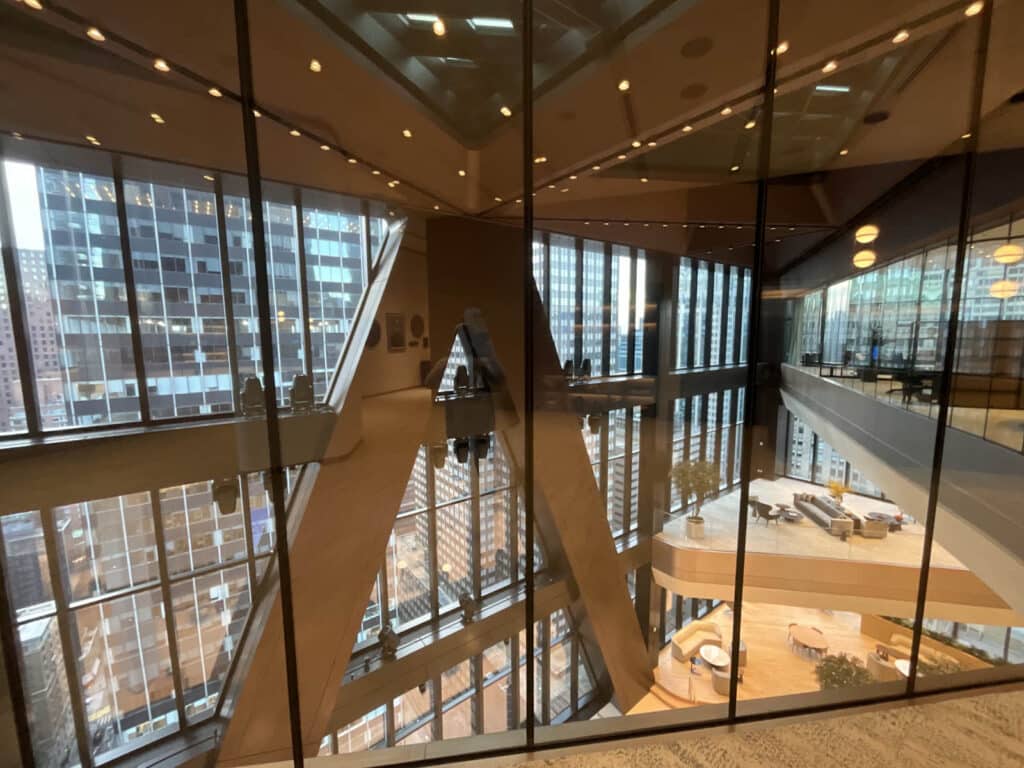 Modern office interior with large glass windows, city skyscrapers reflected in the glass, geometric architectural features, and open lounge areas with tables and chairs visible inside.