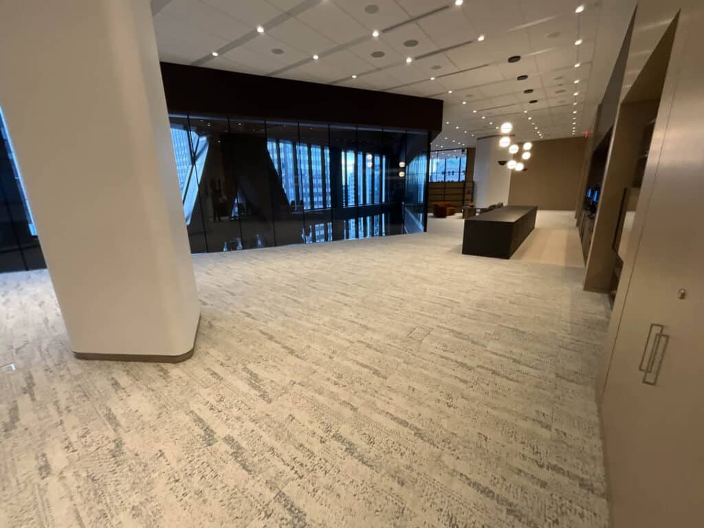 Modern, spacious office lobby with light-colored carpet, high ceiling, globe pendant lights, glass wall overlooking skyscrapers, and a dark reception desk to the right. The space is clean and mostly unfurnished.