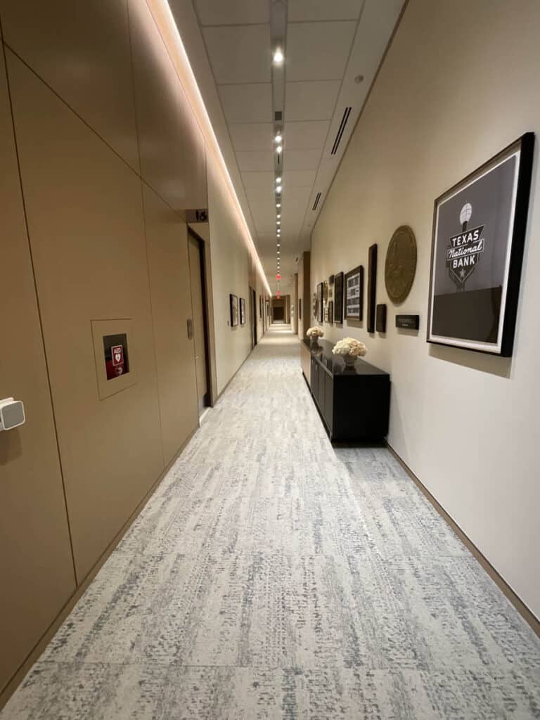 A long, well-lit hallway with beige walls, framed pictures, and plaques on the right. A cabinet with decor and flowers sits along the wall. The light carpet and ceiling create a modern, clean look.