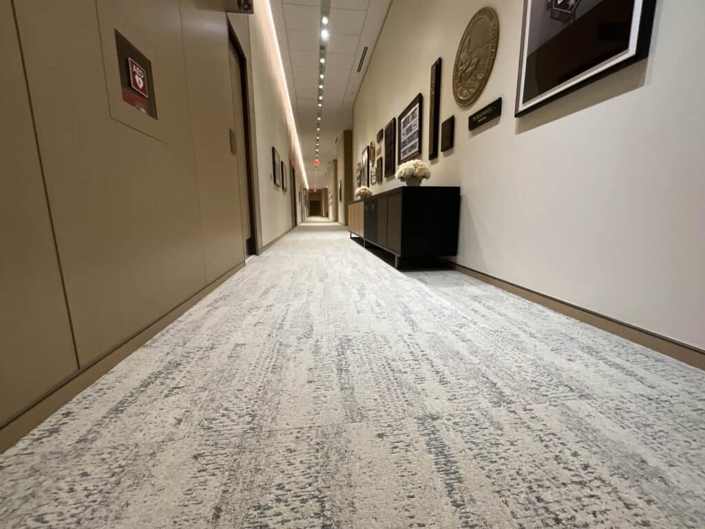 Low-angle view of a hallway with light-colored carpeting, artwork and plaques on the right wall, a black console table, and beige doors on the left side. Ceiling lights illuminate the corridor.