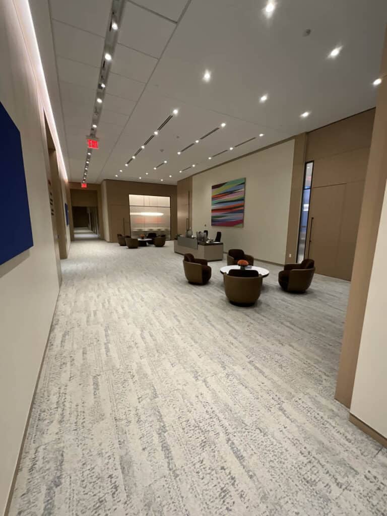 A modern, spacious hallway with beige walls, light carpet, and recessed ceiling lights. Several brown armchairs and round tables are grouped near the end, with colorful abstract art on the walls.