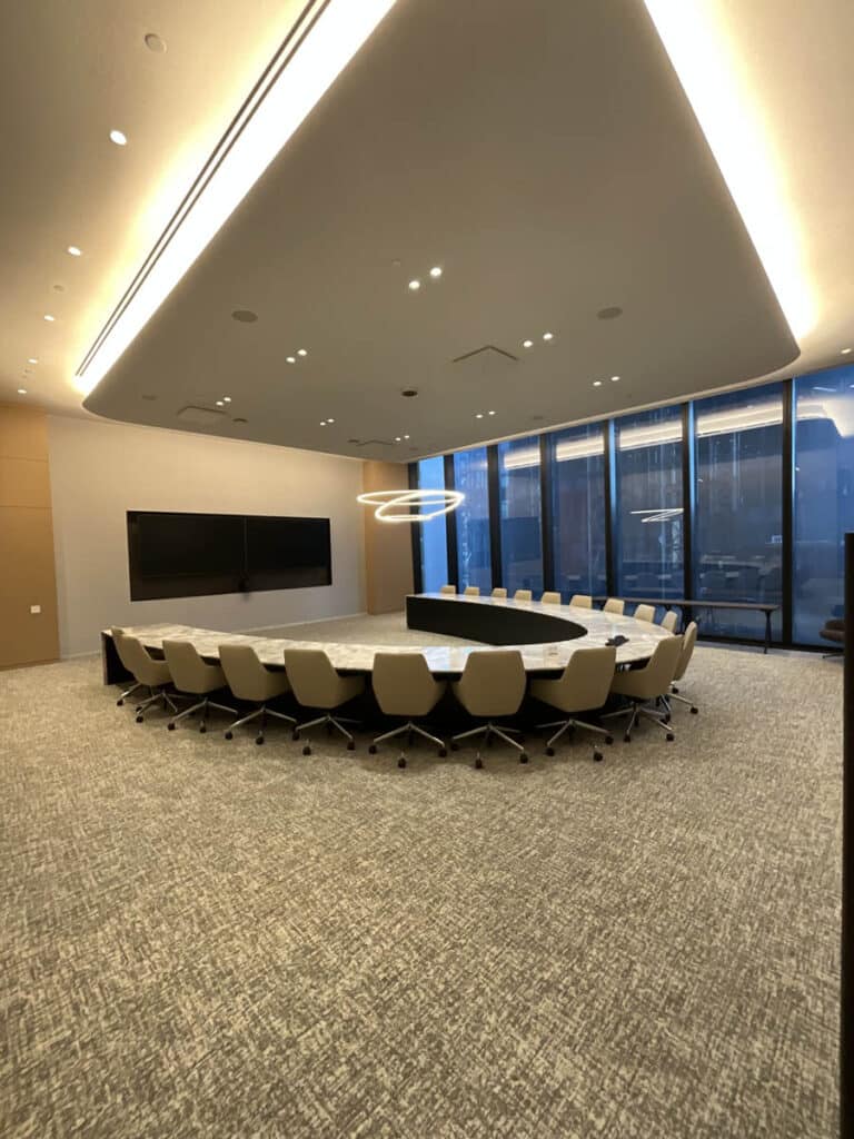A modern conference room with a large, U-shaped table surrounded by beige office chairs, two large wall-mounted screens, floor-to-ceiling windows, and a circular ceiling light fixture.