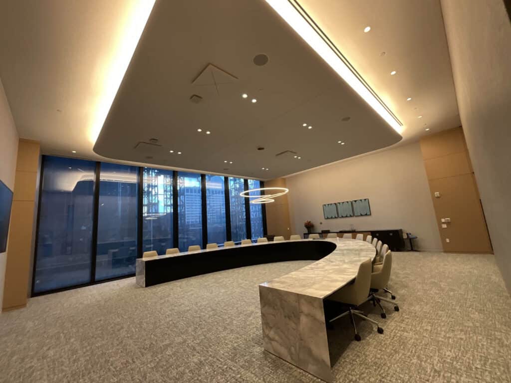 A modern conference room with a large U-shaped marble table, beige chairs, a circular chandelier, floor-to-ceiling windows, and abstract artwork on the wall. The room is spacious and well-lit.