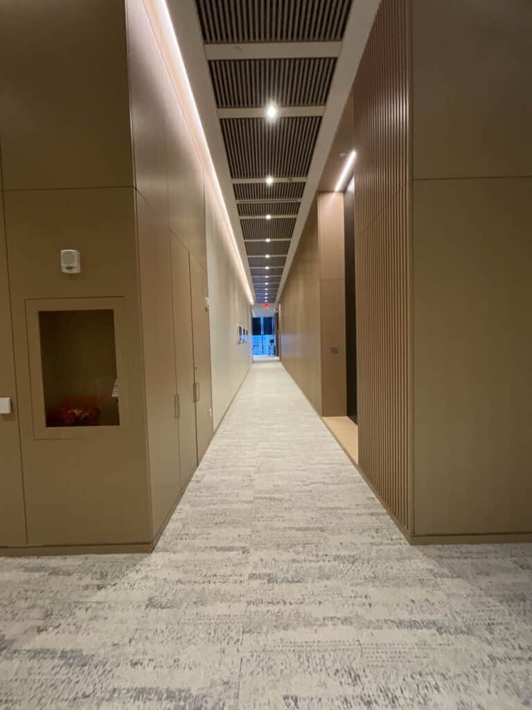 A long, modern hallway with beige walls, a patterned carpet floor, overhead lights, and a ceiling with black slats. The corridor leads to a distant glass door with daylight visible outside.