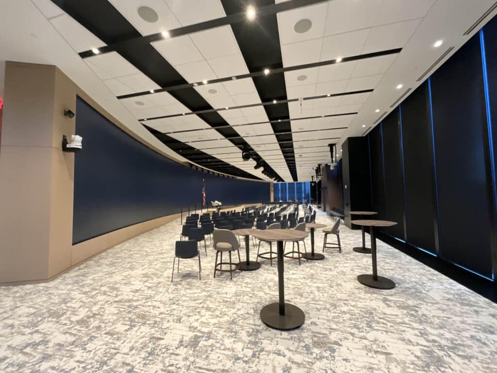 A modern conference room with rows of chairs facing a large screen, round tables with chairs in the foreground, and tall black window shades along one wall. The ceiling has a unique black and white pattern.