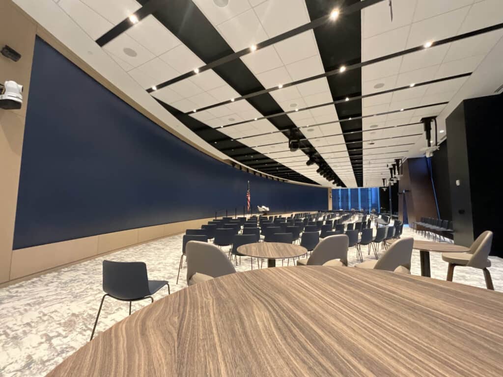 Modern conference room with rows of empty chairs facing a stage with a podium and American flag; large curved blue wall and patterned carpet; photo taken from a wooden table in the foreground.