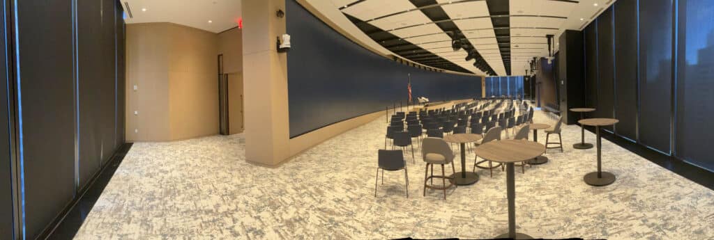 A modern conference room with rows of chairs facing a podium and American flag, round cocktail tables on the right, large windows, and a high ceiling with geometric panels. The room is well-lit and spacious.