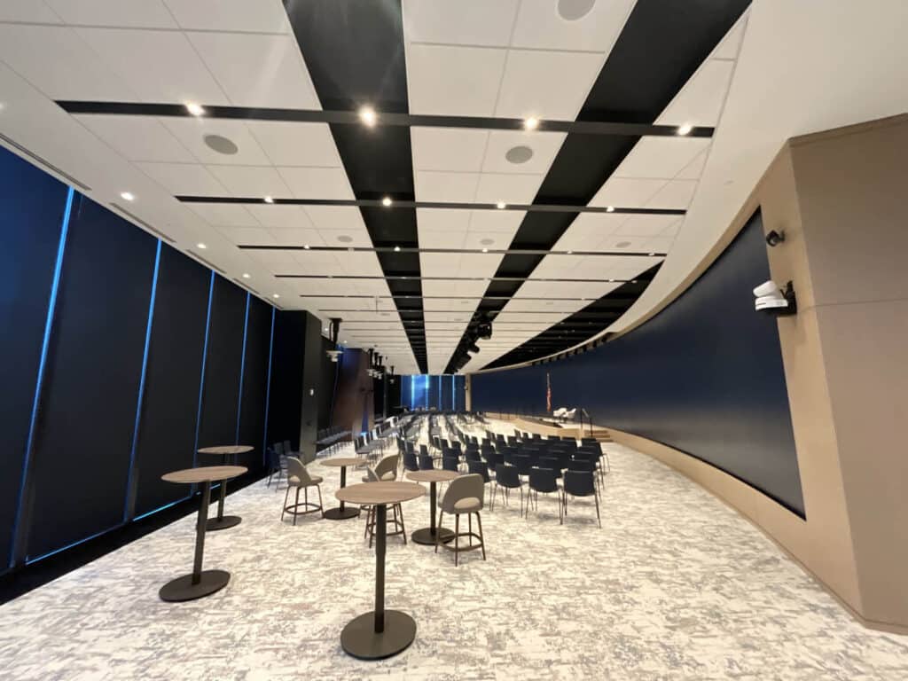 A modern conference room with rows of black chairs facing a stage, small round tables with tall stools on the left, and a curved wall with blue accents on the right under a patterned ceiling.