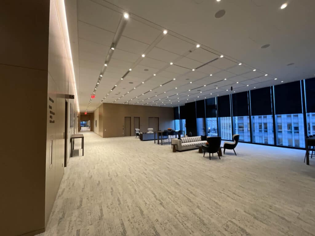 A spacious, modern office lounge with large windows, neutral carpet, sofas, armchairs, and tables. Overhead lights illuminate the open area, which overlooks city buildings outside.