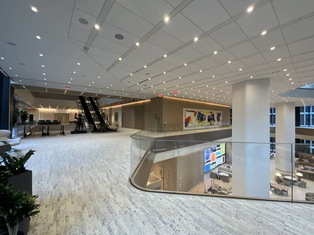 Spacious, modern office lobby with white ceilings, numerous lights, escalators, abstract art on the walls, glass railings, plants, and open areas with seating. Large windows let in natural light.