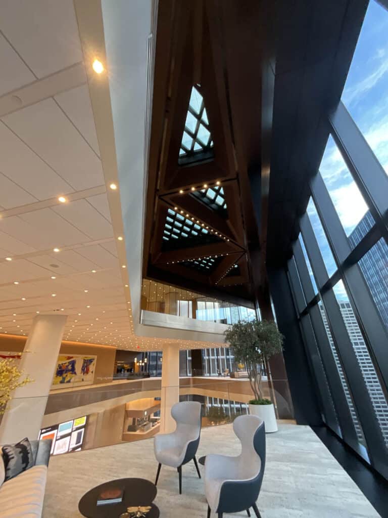 Modern lobby with large windows, high ceilings, geometric lighting, and a mezzanine. Two white chairs and a small table sit near the glass wall, overlooking city buildings outside.