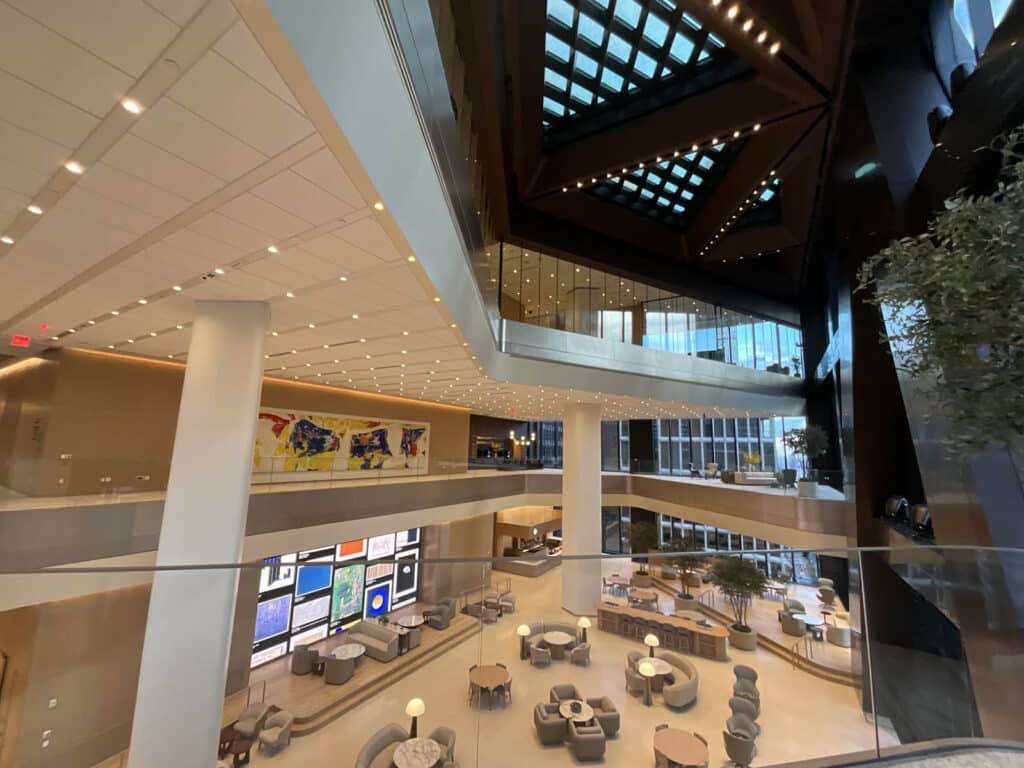 A modern, spacious hotel lobby with multiple floors, large windows, skylights, comfortable seating areas, and vibrant artwork on the walls. Warm lighting and glass railings create an open, inviting atmosphere.